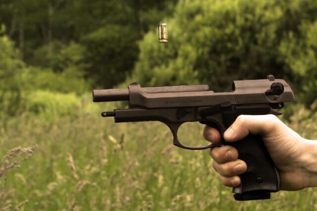 A close-up of a hand illegally possessing a black pistol with a bullet casing ejecting midair